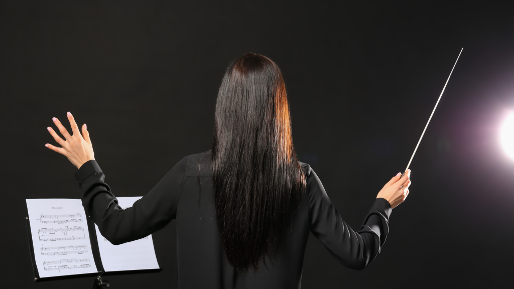 A photo of a brunette music conductor with long hair, arms raised and holding a conductor's baton in her right hand. To her left is a music stand with sheet music on it. This represents tone and pace in voiceover for elearning.